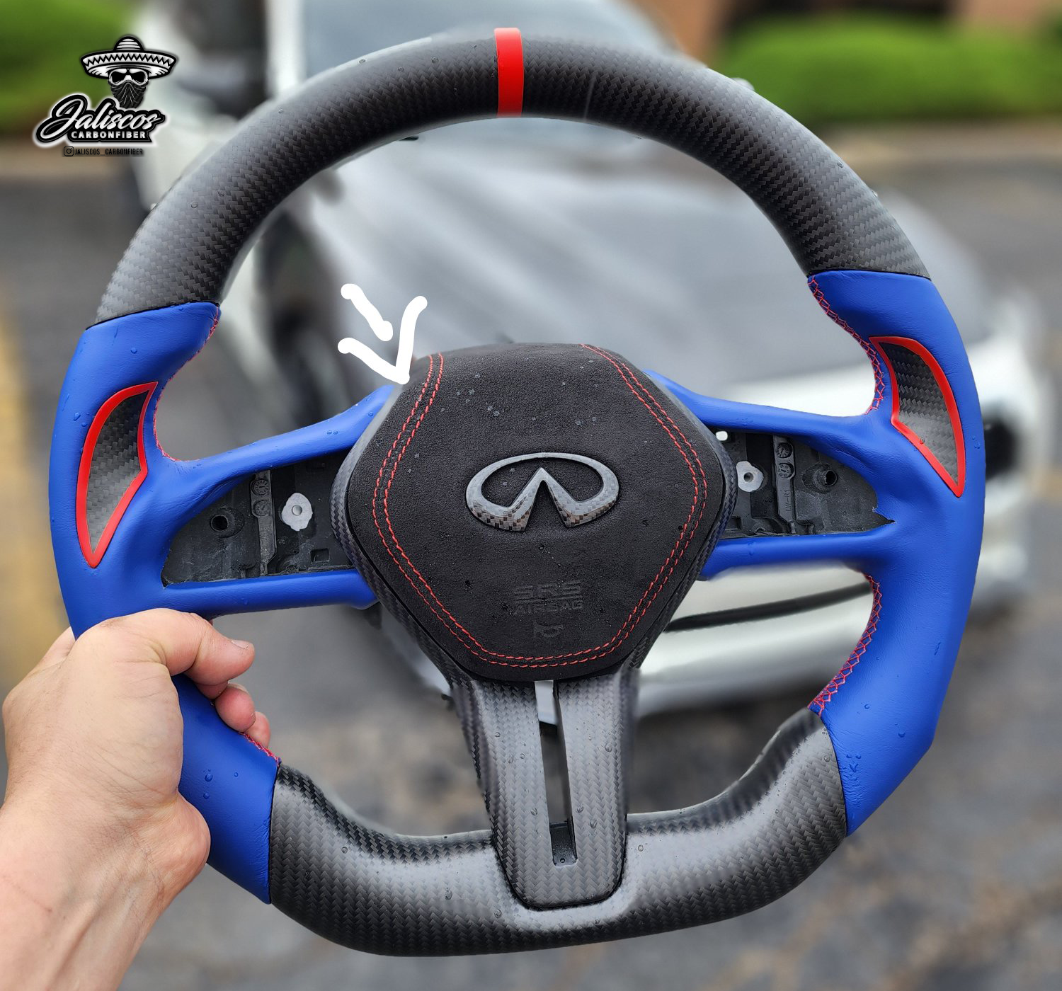 Jalisco's custom cover for Infiniti Q50/Q60 steering wheel airbag: blue grip, black thumb grip with red outlines, red stripes, and a detailed suede-like airbag cover accented with red stitching and a carbon fiber Infiniti logo. Arrow emphasizes the meticulous customization of the airbag cover.
