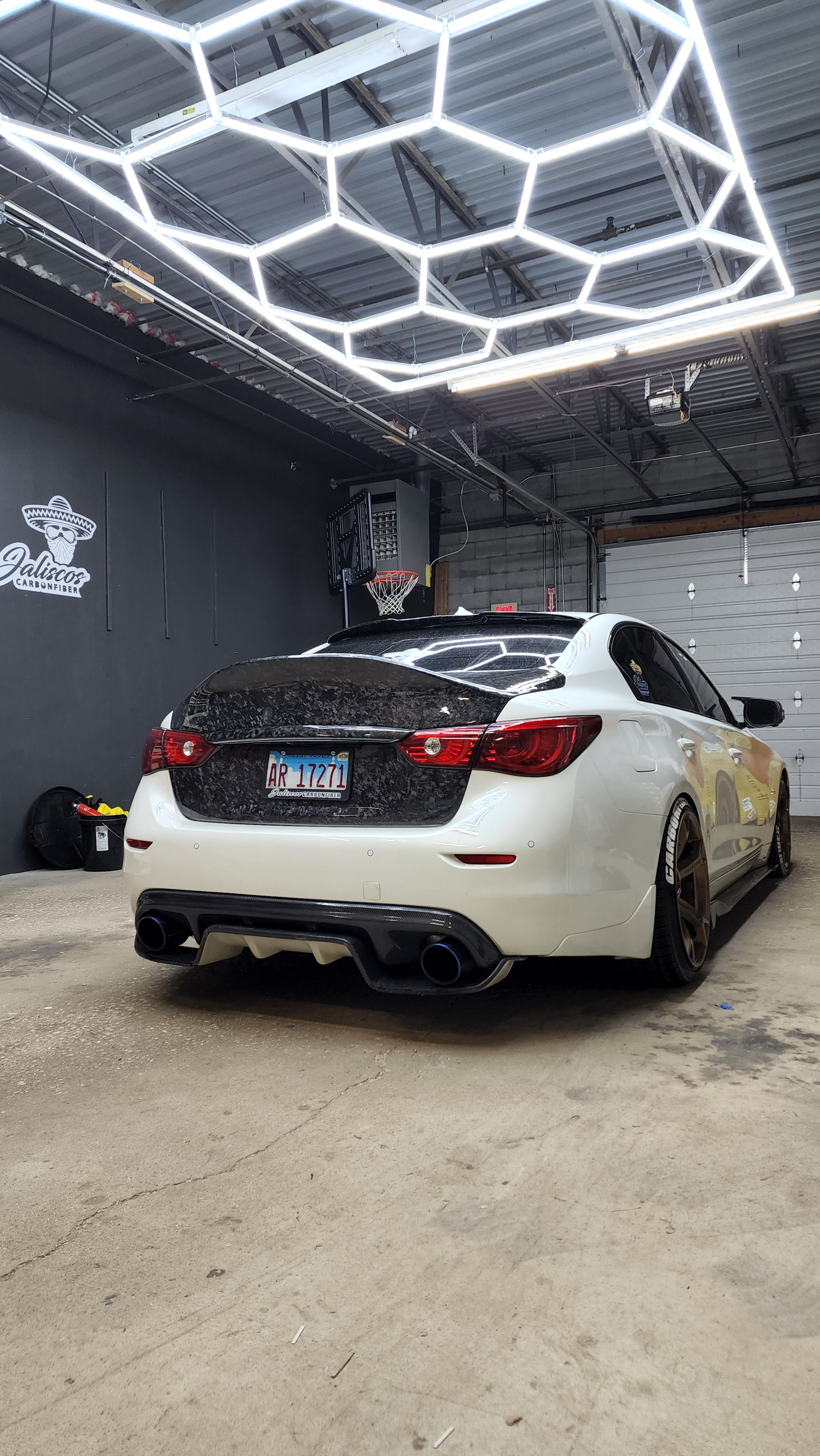 JCF Carbon Fiber Trunk on a white Infiniti Q50 seen from a low angle in a garage, highlighting the trunk's sporty aesthetic and the car's enhanced rear profile.