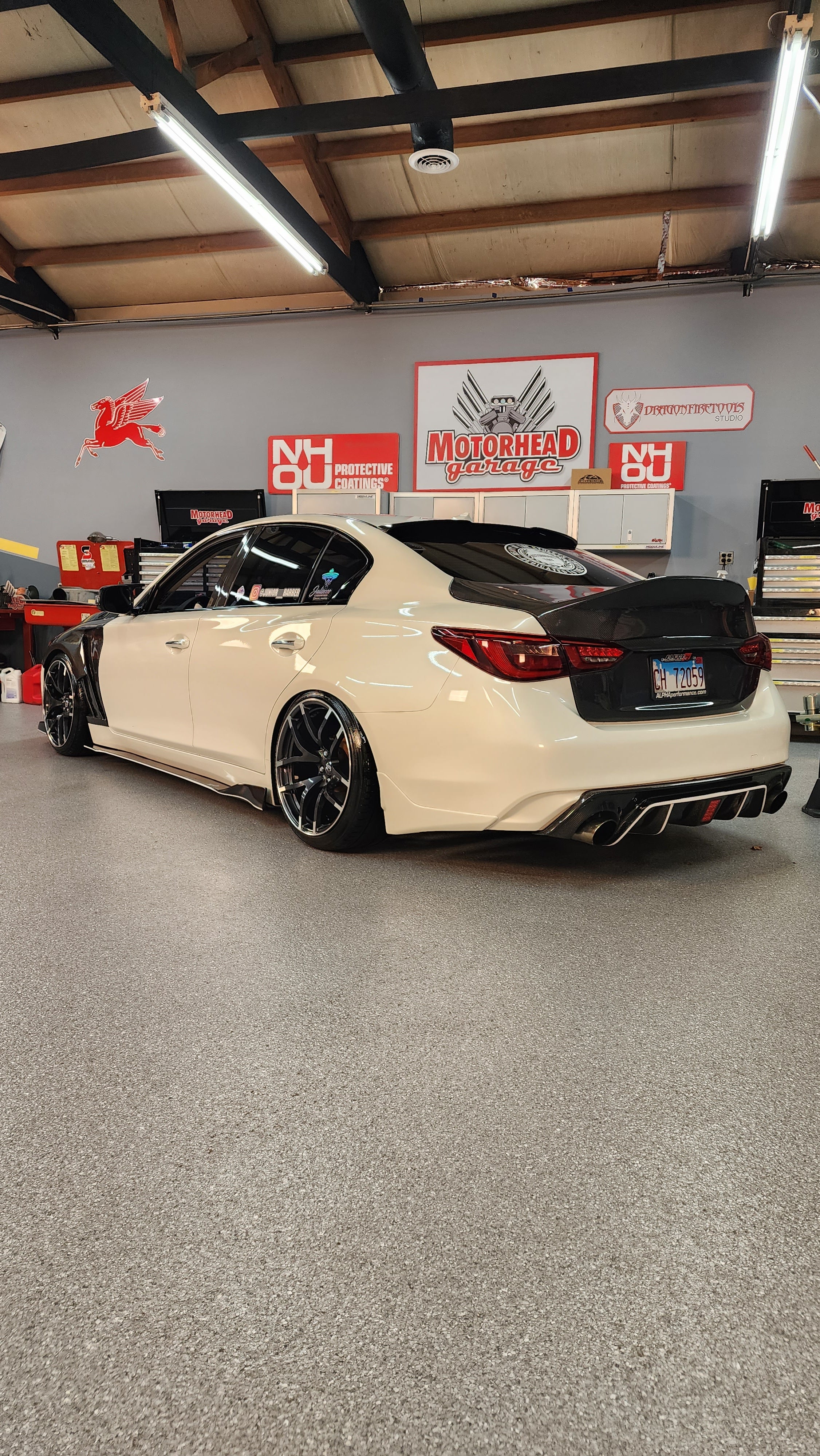 Side rear perspective of a cream-colored Infiniti Q50 with JCF Carbon Fiber Trunk, exhibited in an automotive shop setting, emphasizing the aftermarket upgrade.